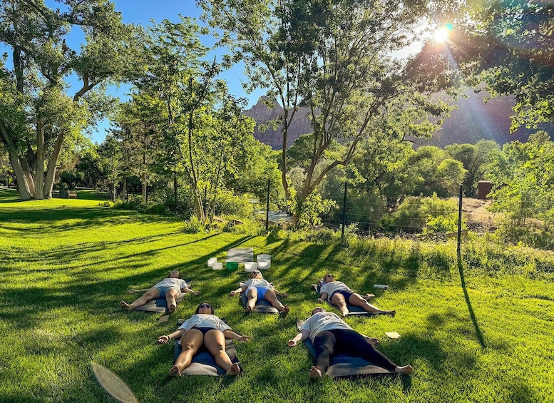 A group of people lying on mats in a grassy park with trees and mountains in the background. 2