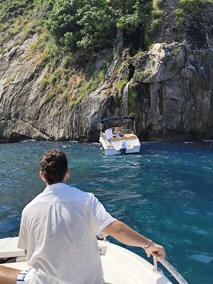 A man steering a boat on clear blue waters, with a rocky cliff and lush greenery in the background. 4