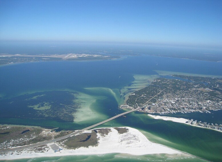 An aerial view of a coastal city with a bridge connecting two land masses, surrounded by a large body of water with varying shades of blue and green, and a sandy beach area. 1