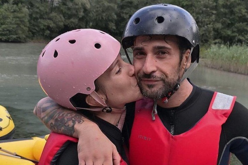 Two people wearing helmets and life jackets, one in a pink helmet and the other in a black helmet, with a river and greenery in the background. 2