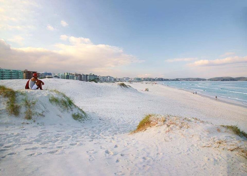 A beach with sand dunes, a person sitting on a dune, and buildings in the background. 5