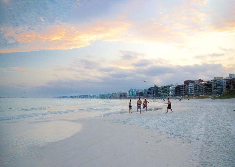 A beach with people walking and playing, buildings in the background, and a sunset sky. 3