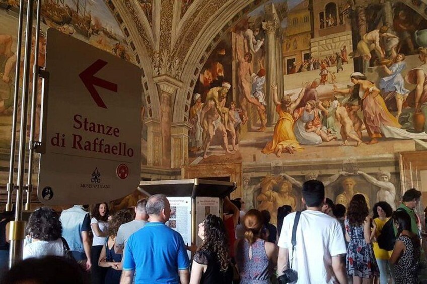 A crowded museum with a large fresco mural on the wall and a sign pointing to the Stanza di Raffaello. 4