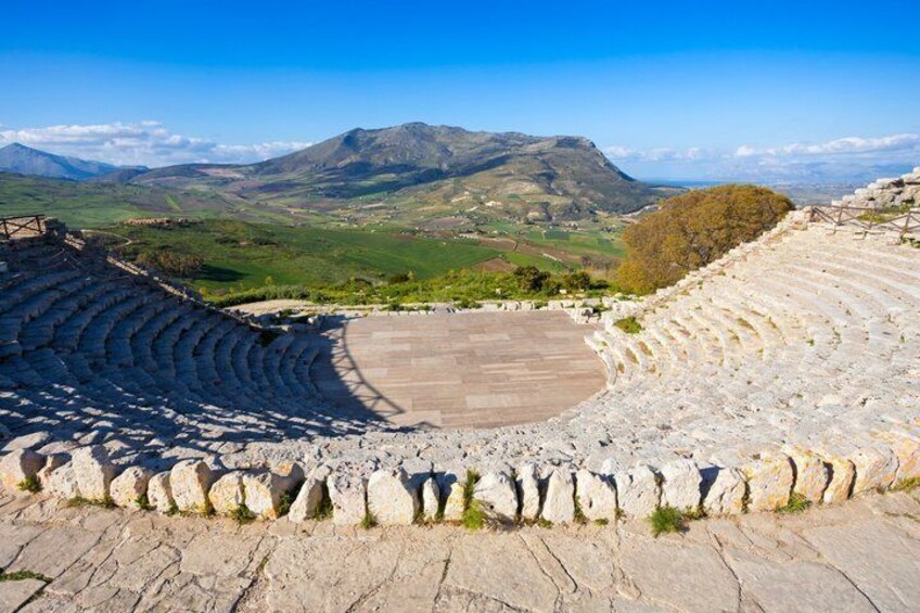 An ancient amphitheater with stone seating and a central stage, surrounded by a mountainous landscape. 3