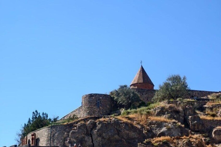 A historic stone fortress with a conical roof and a round tower, surrounded by rocky terrain and sparse vegetation. 5