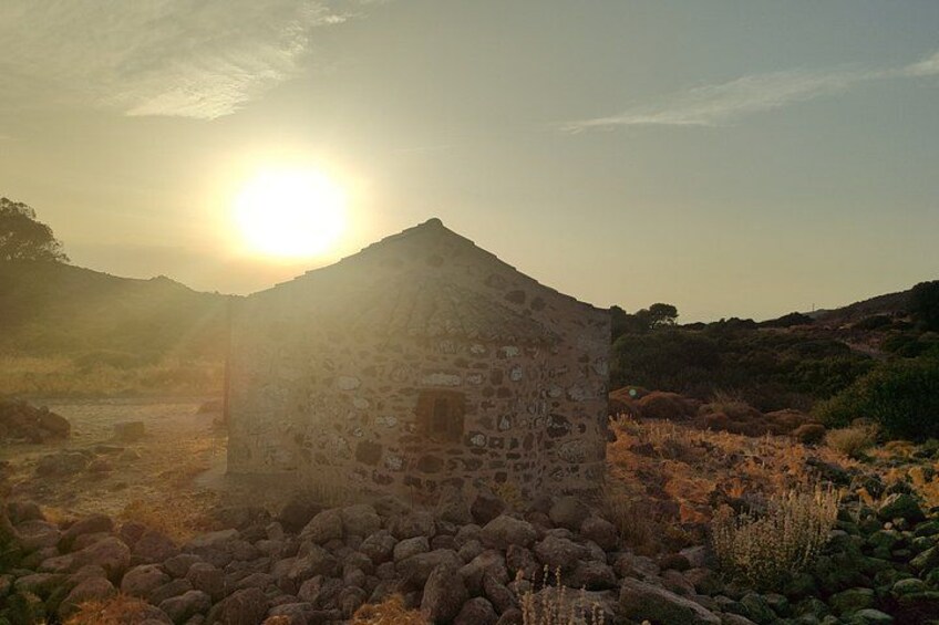 A stone building with a sloped roof, surrounded by dry grass and rocks, with the sun low in the sky. 3