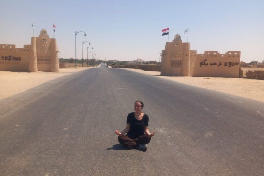 A person sitting on the road with a desert landscape and two gates with flags in the background. 3
