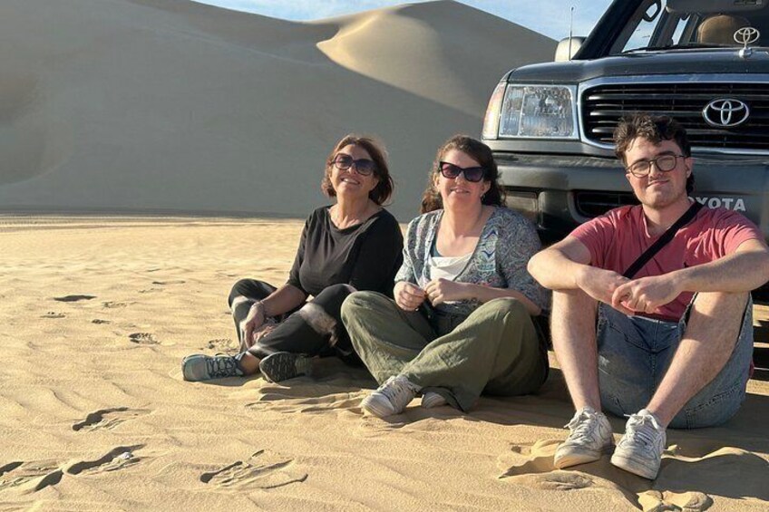 Three people sitting on sand dunes with a Toyota vehicle in the background. 1