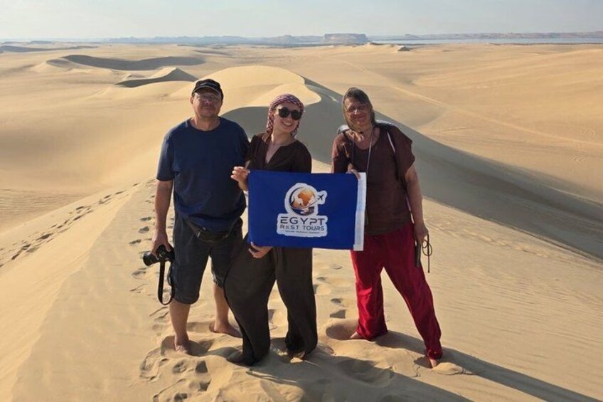 Three people standing on a sand dune with a logo on a board. 2