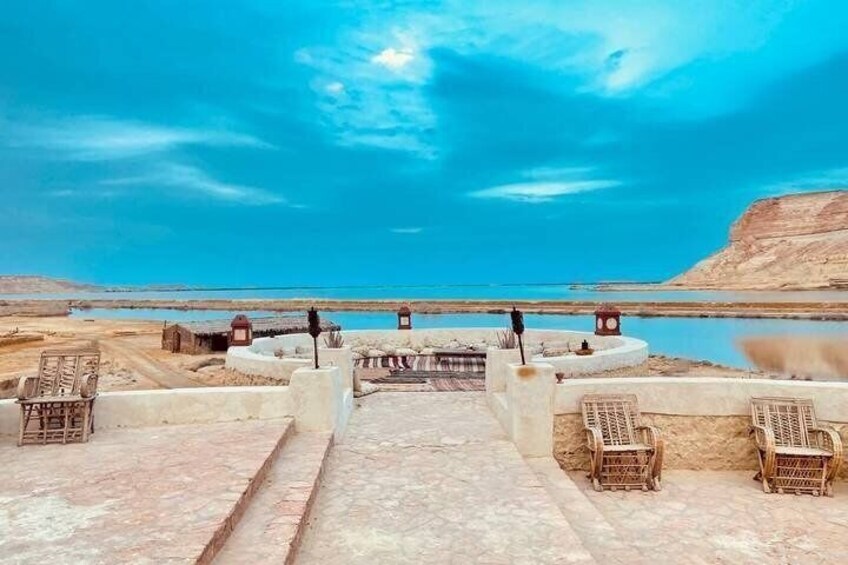 A coastal landscape with a clear sky, a calm sea, and a series of steps leading to a viewing platform with wicker chairs. 2