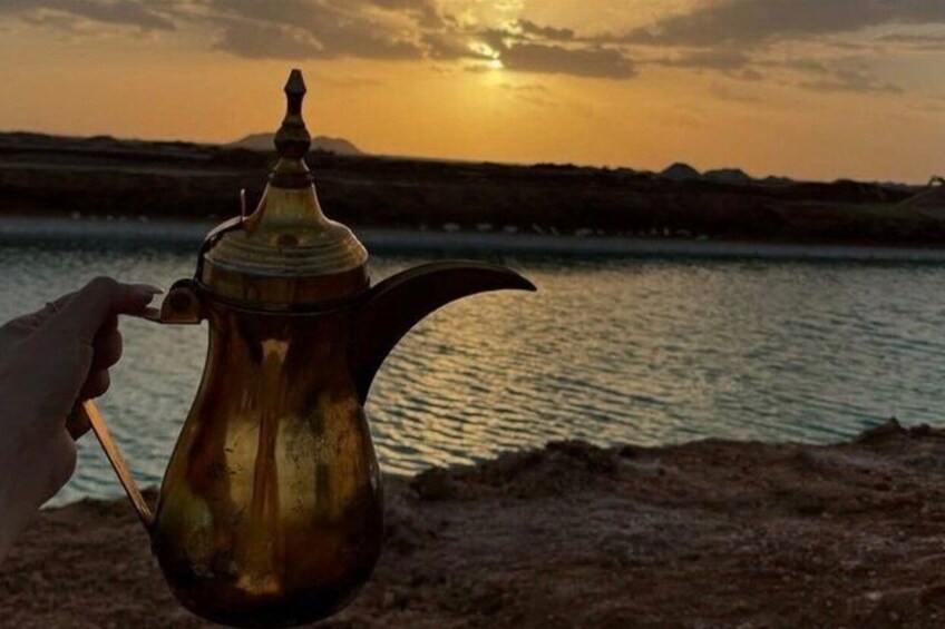 A hand holding a brass teapot with a spout and handle, set against a sunset over a body of water. 5