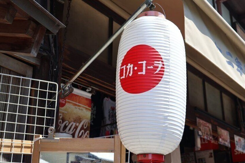 A traditional Japanese paper lantern with red and white colors, attached to a metal stand, in front of a building with a visible Coca-Cola sign. 2
