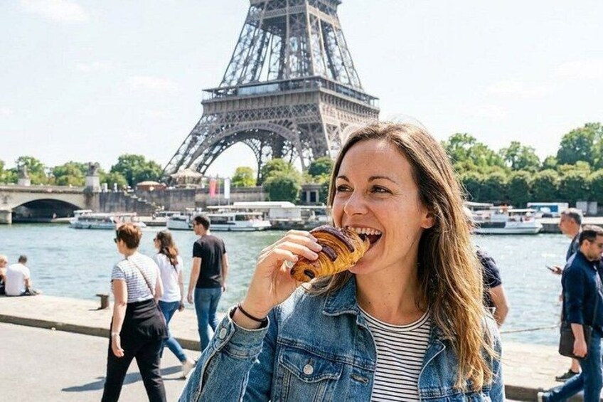 A person eating a pastry near the Eiffel Tower with the Seine River in the background. 1