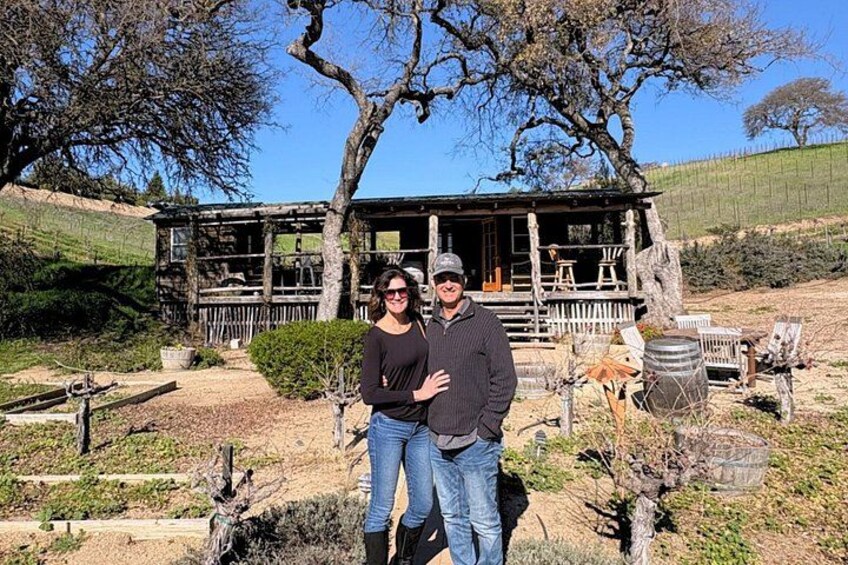 A wooden cabin with a porch, surrounded by a vineyard and a large tree. 2