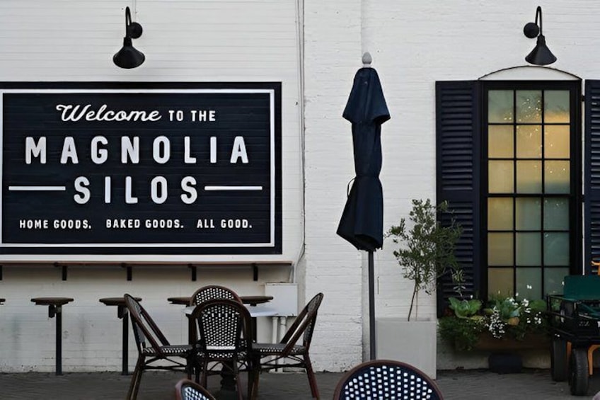 Ein Geschäft mit einer Schaufensterschild, auf dem 'Welcome to the Magnolia Silos' steht und Haushaltswaren sowie Backwaren bewirbt. Vor dem Geschäft befinden sich Tische und Stühle im Freien. 1