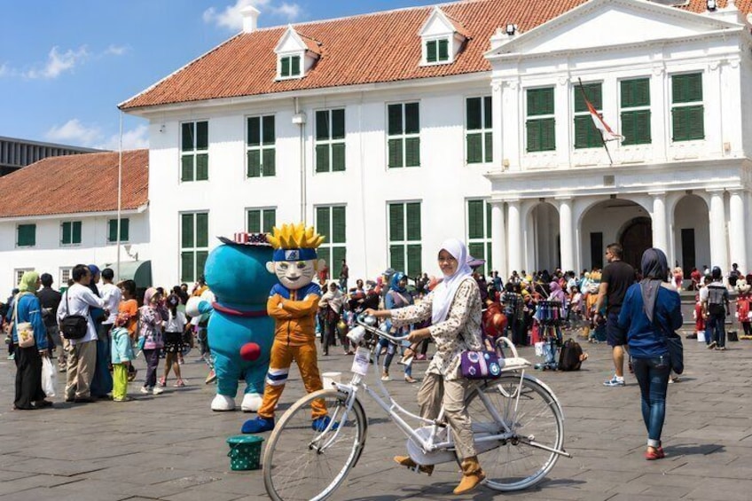 A public square with a white building with green shutters, a person on a bicycle, and a character in a blue costume. 1