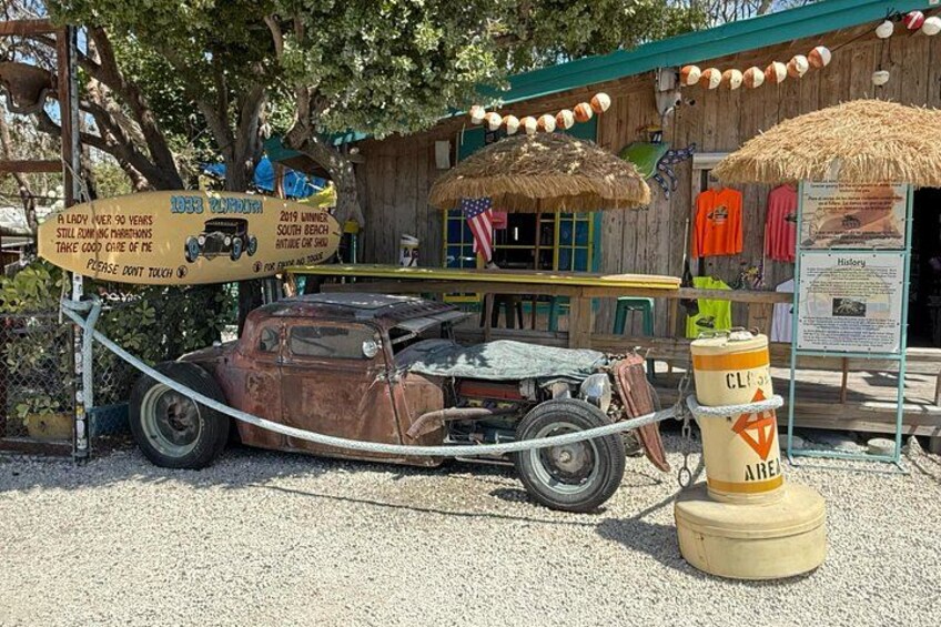 A vintage car on display, tied to a post with a rope, in front of a wooden building with a sign. 4
