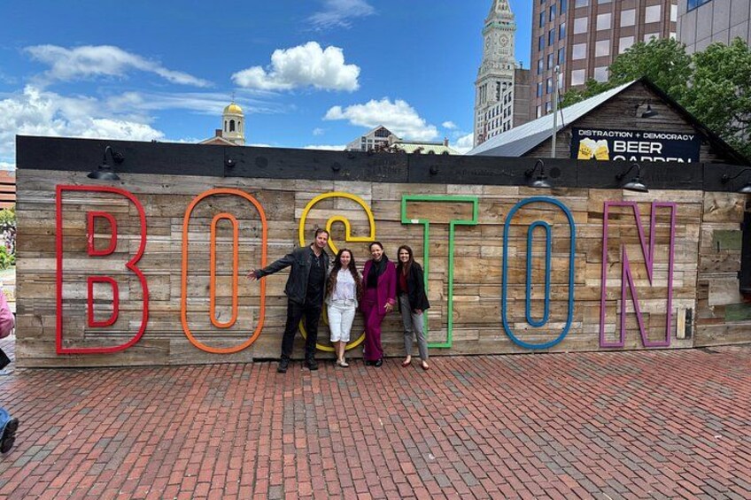 A wooden sign with the word 'BOSTON' in colorful letters, a brick pavement, and buildings in the background. 4
