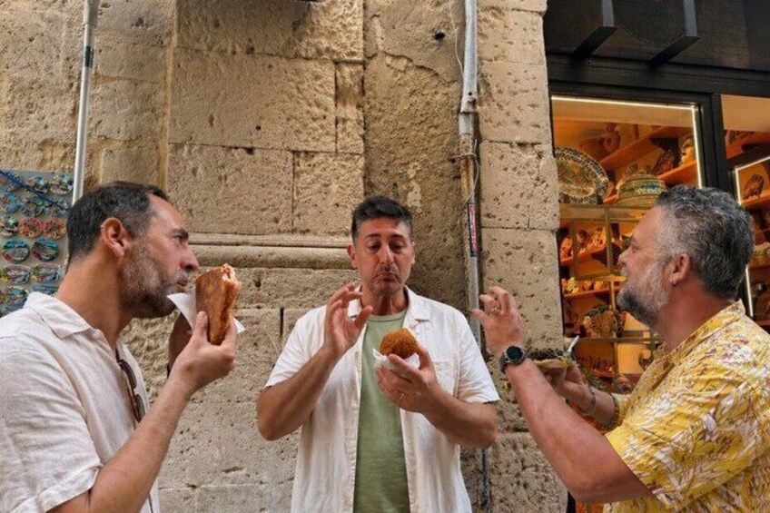 Three men eating pastries in front of a building with a textured wall. 2
