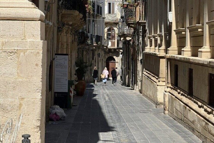 A narrow street with buildings on either side, a signboard, and a few pedestrians walking. 4