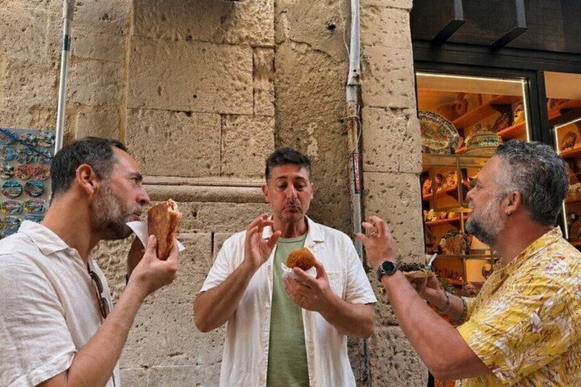 Three men eating food in front of a building with a textured wall and a shop with colorful items in the background. 3
