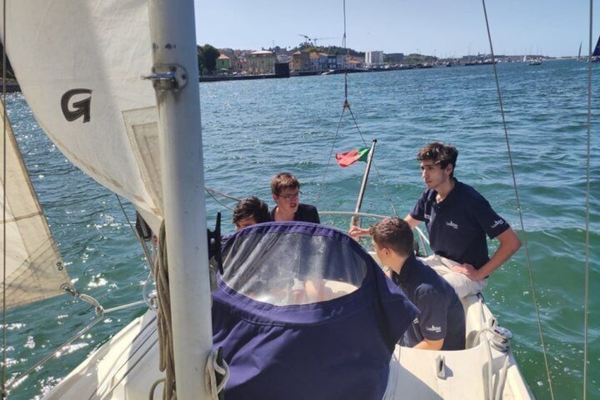 A sailboat with a person steering and others on board, water in the background, and a distant shoreline with buildings. 5