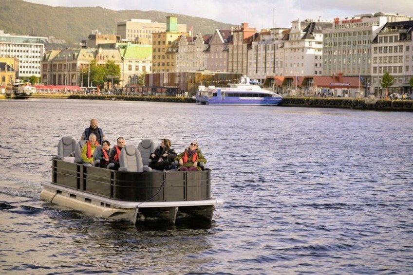 A boat with passengers on a river, a larger ship in the background, and buildings along the riverbank. 3