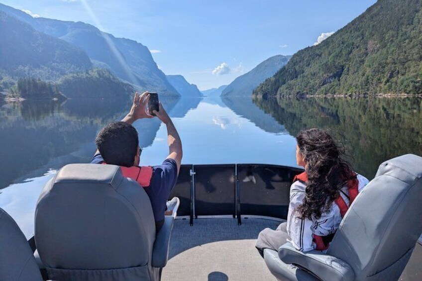 Two people on a boat, one taking a photo, with a calm lake and mountains in the background. 1