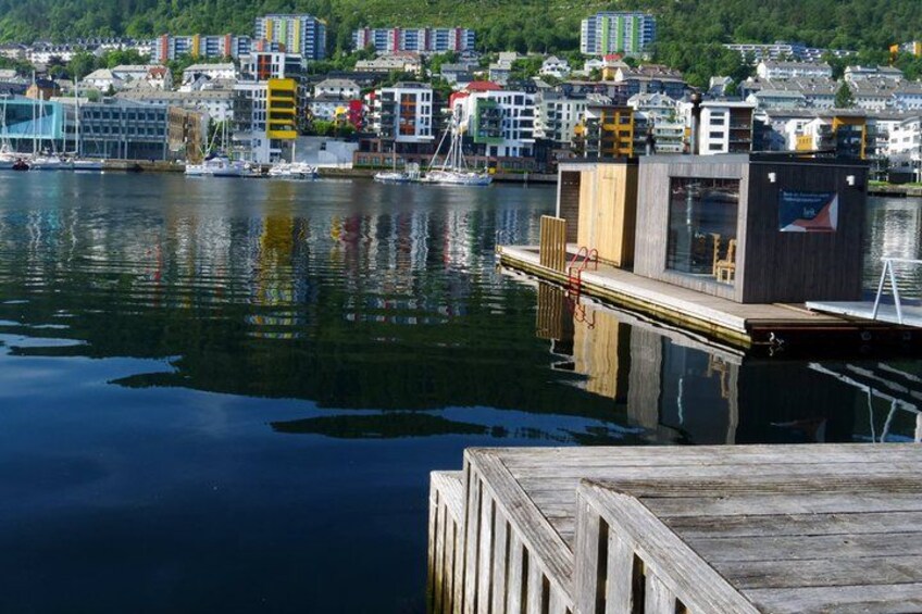 A waterfront with a wooden dock, a small building, and a cityscape in the background. 2