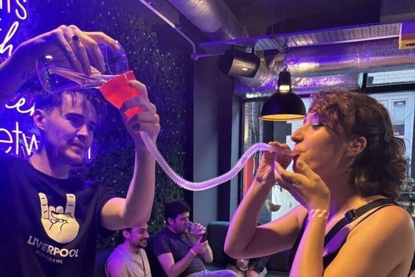A person holding a glass with a hose, another person drinking from a straw, and a neon sign in the background. 1