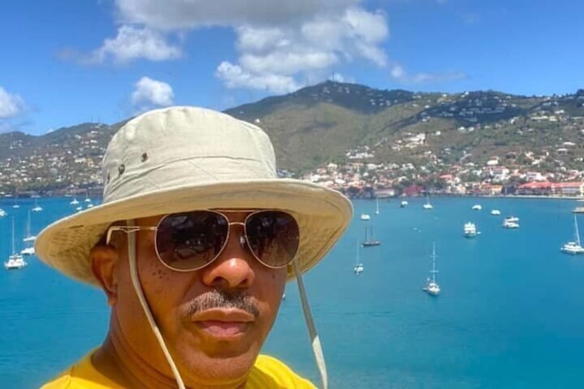 A person in a yellow shirt and white hat with a coastal town and boats in the background. 4