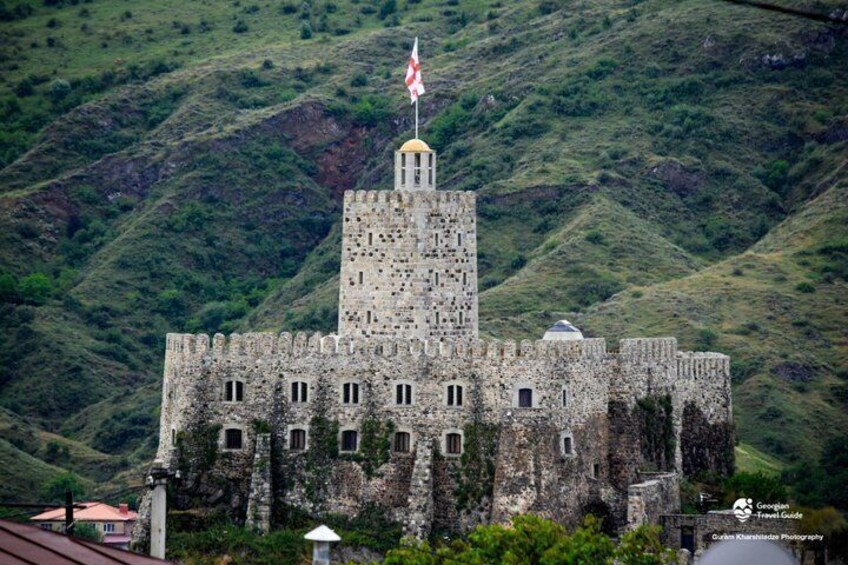 A historic stone castle with a flag on top, surrounded by lush green hills. 2