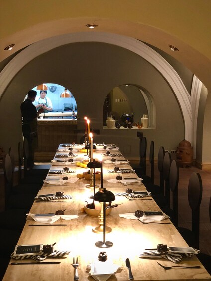 A long dining table set with candles, plates, and cutlery, with a chef in the background. 2