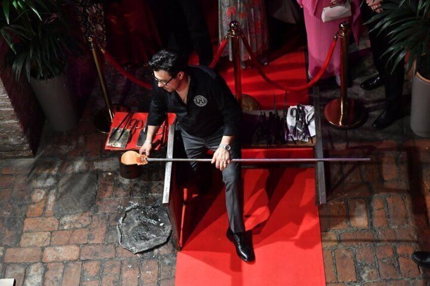 A person sitting on a red carpeted bench with a guitar, surrounded by potted plants and a red rope barrier. 3