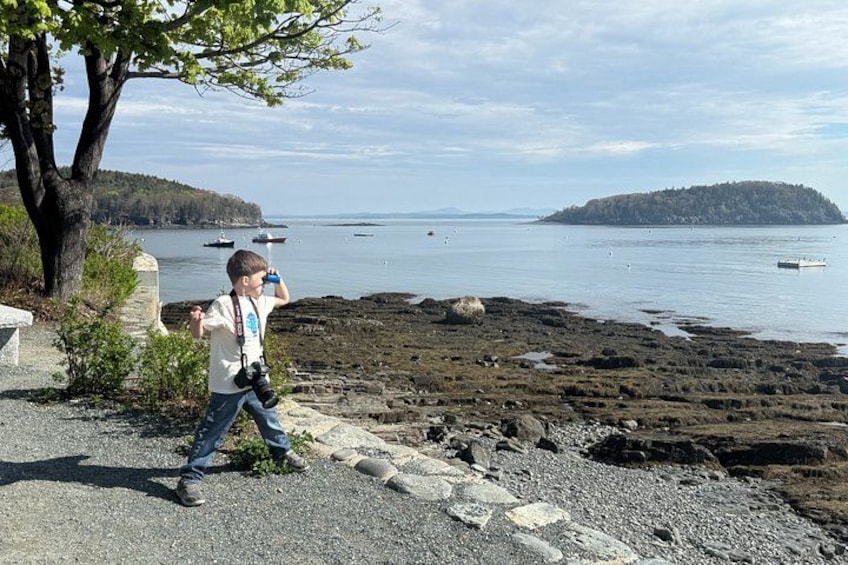 A rocky shoreline with a person taking a photo, boats in the water, and a distant island. 1