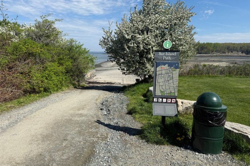 A gravel path leading to Bar Island Path with a map sign, surrounded by greenery and a clear sky. 2
