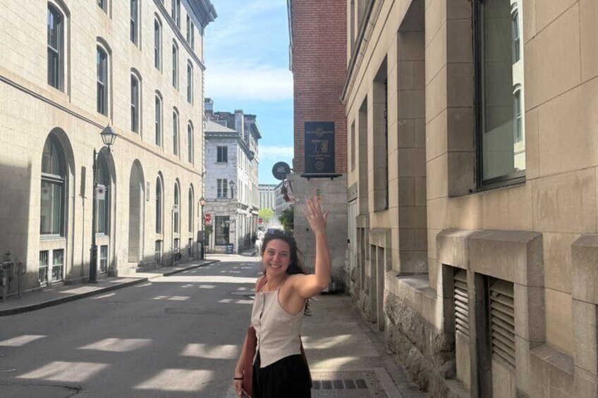 A woman standing on a street with historic buildings, a clear sky, and a signboard on the right. 2