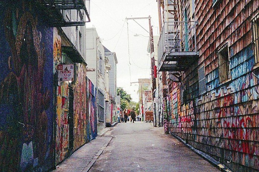 A narrow alley with graffiti-covered walls, a sidewalk, and a few pedestrians in the distance. 1