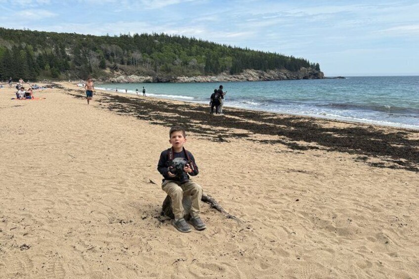 A beach with people, a child with a camera, and a forested coastline. 5