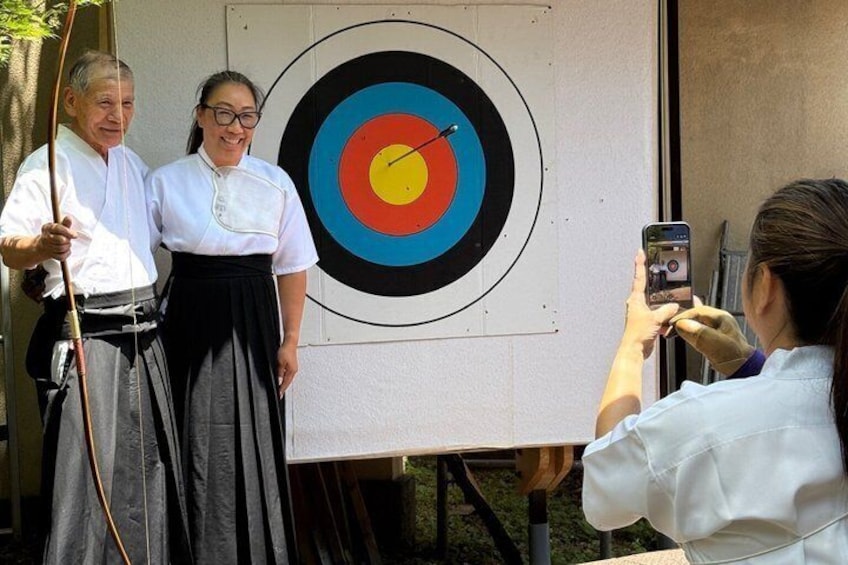 An outdoor archery setup with a target, an archer, and a person taking a photo. 2
