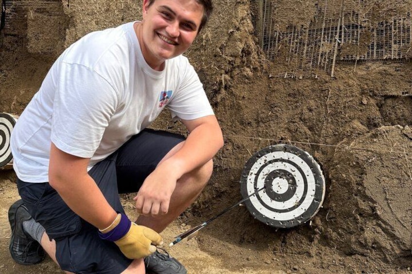 A person crouching next to a target with concentric circles, in a sandy outdoor setting. 5