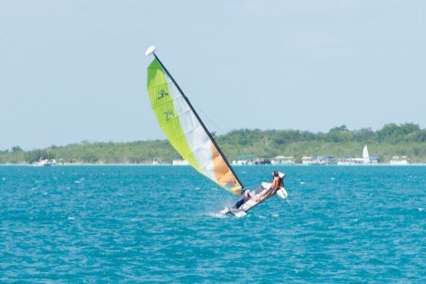 A person windsurfing on a body of water with a green sail, surrounded by a calm sea and a distant shoreline with buildings. 5