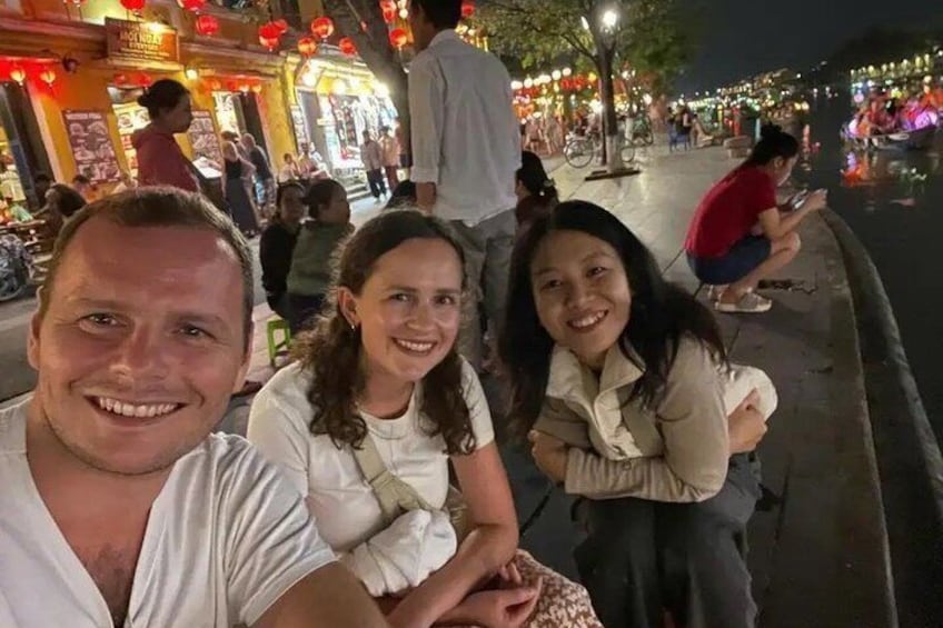 A group of people sitting on a bench in an outdoor area with shops and street lamps in the background. 3