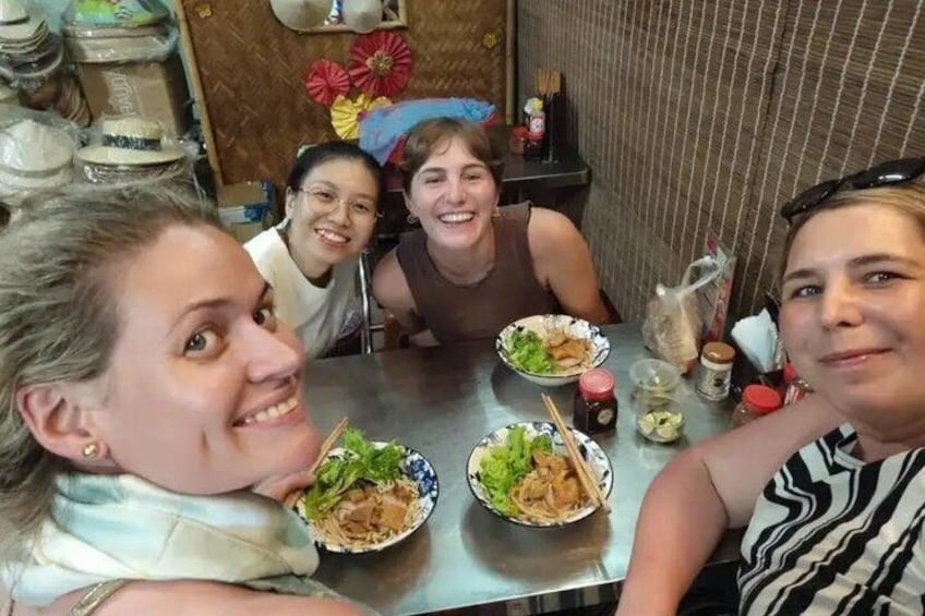 A group of people sitting at a table with bowls of food, a metal counter, and various kitchen items in the background. 2