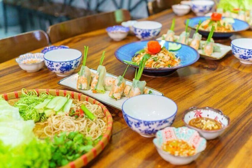 A wooden table with various dishes, including noodles, spring rolls, and bowls of dipping sauces. 1