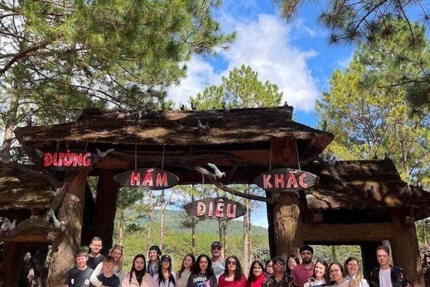 A group of people posing in front of a rustic wooden structure with signs reading 'DUONG,' 'HAM,' 'KHAC,' and 'DIEU.' 4