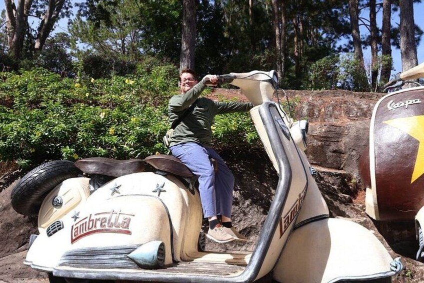 A person on a vintage vehicle with a logo, surrounded by greenery and trees. 1