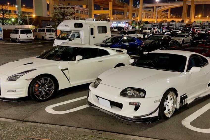 Two white sports cars parked in a lot with a large RV and other vehicles in the background. 3