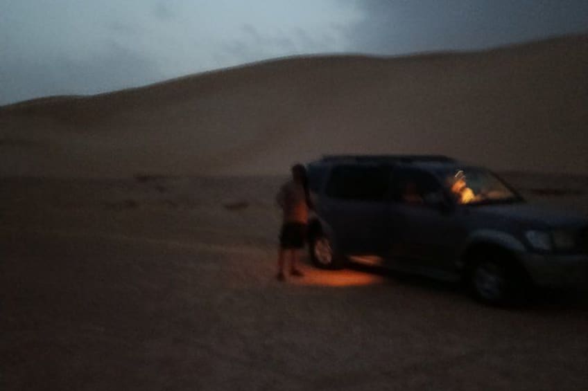 A person standing next to a vehicle in a desert with sand dunes in the background. 5