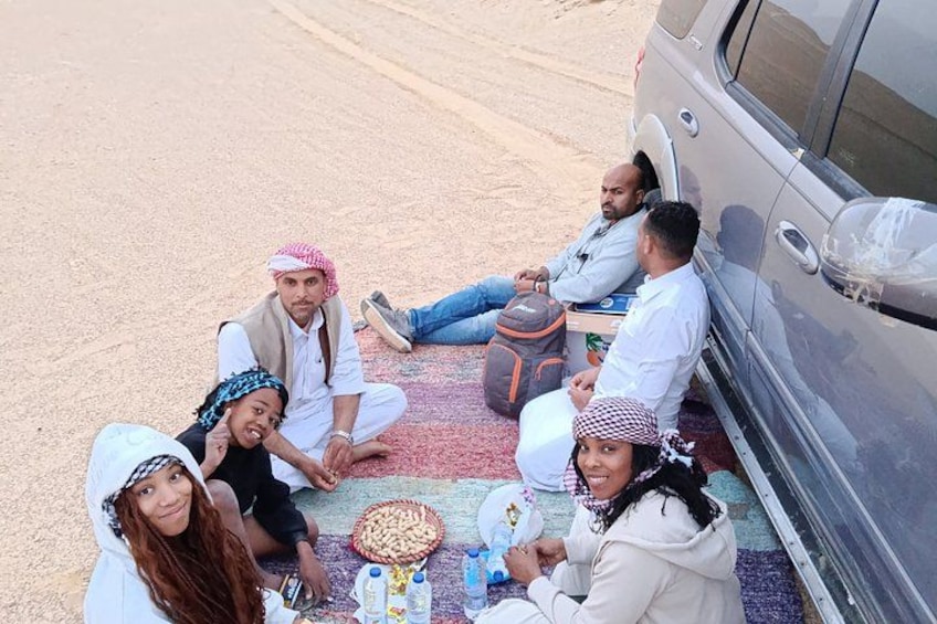 A group of people sitting on the ground around a vehicle, with a carpet and some items on the ground. 4